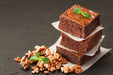 Chocolate brownie square pieces in stack on white plate with walnuts, decorated with mint leaves and cocoa on black background. Delicious dessert. Dark mood. Close up photography. Selective focus