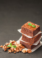 Chocolate brownie square pieces in stack on white plate with walnuts, decorated with mint leaves and cocoa on black background. Delicious dessert. Dark mood. Close up photography. Selective focus