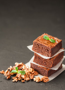 Chocolate brownie square pieces in stack on white plate with walnuts, decorated with mint leaves and cocoa on black background. Delicious dessert. Dark mood. Close up photography. Selective focus