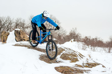 Cyclist in Blue Riding Mountain Bike on Rocky Winter Hill Covered with Snow. Extreme Sport and Enduro Biking Concept.