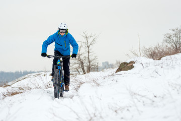 Fototapeta na wymiar Cyclist in Blue Riding Mountain Bike on Rocky Winter Hill Covered with Snow. Extreme Sport and Enduro Biking Concept.