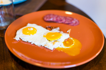 Closeup of two broken runny fried eggs on large orange plate with cured meat hard salami slices