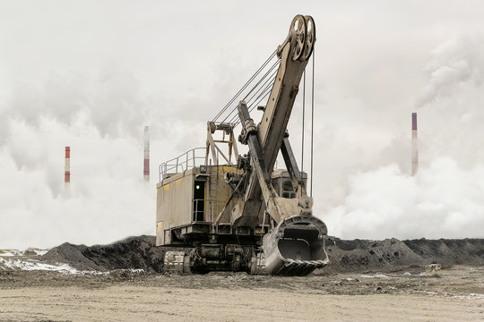 Heavy bucket mining excavator on a crawler track against a background of dense factory strong smog with smoking chimneys. Excavator with mechanical drive and flexible suspension of work equipment.