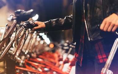 Young girl in black leather jacket using bike on background illumination glow bokeh light in night atmospheric city, hipster biking and riding to work by bicycle on urban street, ecology concept.