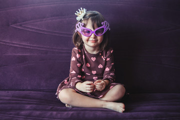 Portrait of cute adorable little girl with funny glasses sitting on couch indoors. Preschooler having fun at home. Lifestyle happy childhood concept. Kid celebrating holiday