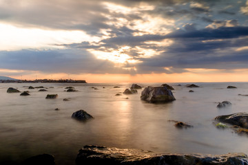 sun rays over the black sea and the cloud sky. Rocks in the water. Long exposition