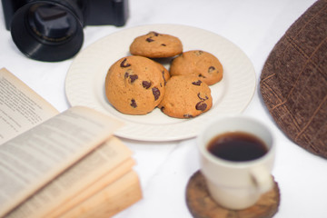 Table with chocolate chips cookies, book, cup of coofee, camera and hat