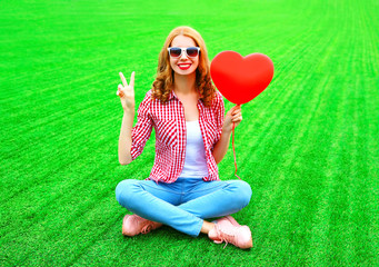 Portrait smiling woman holds red an air balloon in the shape of a heart sits on the grass