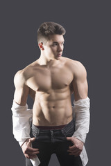 Fototapeta premium Handsome young muscular man shirtless wearing jeans, taking off leather jacket on naked muscle torso, on dark background in studio shot