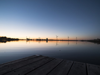 Ocean/lake after sunset with industrial buildings and windmils in the background and a clear blue and orange sky
