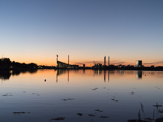 Ocean/lake after sunset with industrial buildings and windmils in the background and a clear blue and orange sky