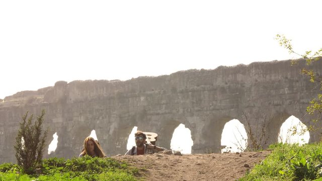 Young lovely couple backpackers tourists walking holding hands toward roman aqueduct arches in parco degli acquedotti park ruins in rome on romantic misty sunrise with guitar and sleeping bag slow