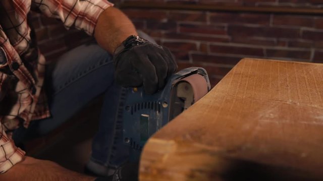 close up shot of the man's hands in protective gloves, who holds an electric grinder to work with a wooden table, the person carefully handles the edges of the table