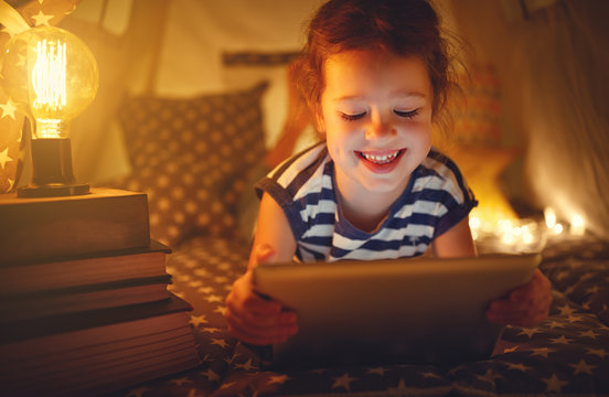 happy child girl with tablet pc laughing in dark in tent at home