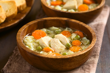 Hühnersuppe mit Suppennudeln, Erbsen und Karotten in Holzschüssel, fotografiert mit natürlichem Licht (Selektiver Fokus, Fokus auf das vordere Fleischstück in der Suppe)