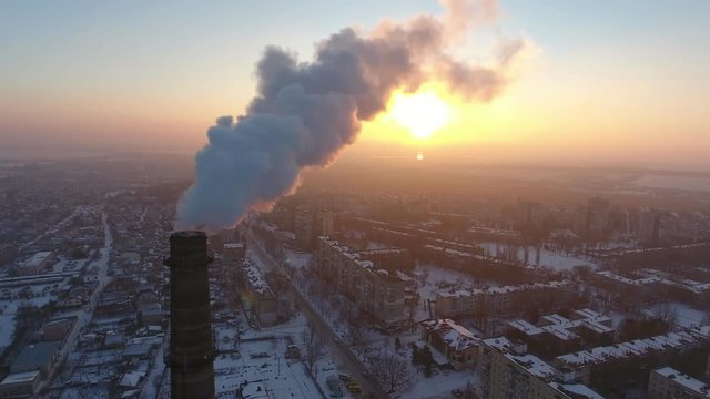 A picturesque bird`s eye view of a high brick chimney with a slow flow of white smoke at a splendid sunset in winter. The cityscape is fine.
