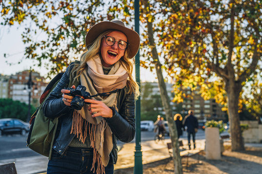 Sunny day, autumn.Young woman tourist, photographer, hipster girl dressed in hat and eyeglasses,sits on bench on city street and takes photo.Vacation, travel,adventure, sightseeing.Blurred background.
