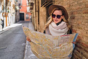 Tourist girl in hat and sunglasses stands on city street near stone wall and looking for way on map. Young attractive woman standing and holding map. Blurred background.