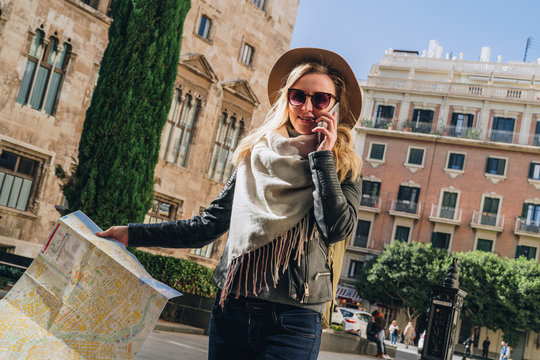 Young smiling woman tourist in hat and sunglasses stands on street of ancient European city and holds map in her hands. In background is beautiful building. Tourism, travel, vacation, trip, journey.