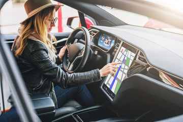 Young woman sits behind wheel in car and uses an electronic dashboard, tablet computer. Girl is...