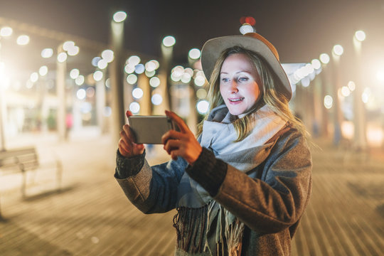 Night. Young smiling woman, wearing in hat and coat is standing on city street and is looking at screen of smartphone in her hands. Girl uses digital gadget, chatting, blogging, checking email.