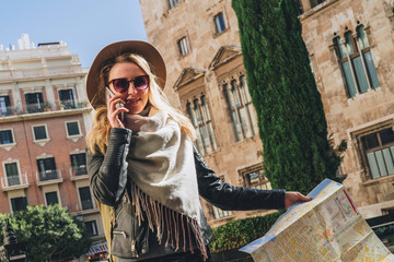 Young smiling woman tourist in hat and sunglasses stands on street of ancient European city and holds map in her hands. In background is beautiful building. Tourism, travel, vacation, trip, journey.