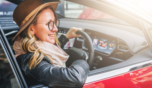 Side view, close up. Young smiling woman in glasses and hat sits behind wheel in red car. Trip, caravanning, tourism, journey.
