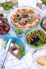 Round large pizza from the oven with prosciutto and olives, arugula and basil. Table setting for lunch or dinner. Table and dishes in blue.