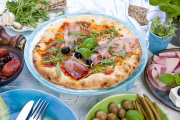 Round large pizza from the oven with prosciutto and olives, arugula and basil. Table setting for lunch or dinner. Table and dishes in blue.