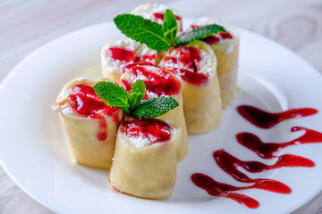 Pancakes stuffed with cottage cheese on a white plate, poured with jam and decorated with green mint leaves.