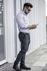 Handsome trendy man wearing white shirt standing and typing on cell phone, outdoor in city setting in day shot