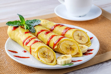 On a light table, a white plate with bananas wrapped in pancakes. Pancakes are poured with jam and decorated with green mint leaves. Near the plate are a white saucer and a cup.