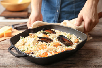 Woman with frying pan of delicious seafood risotto at wooden table