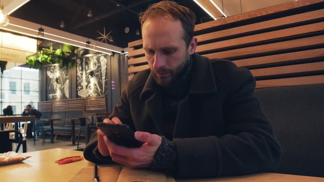 4k Breaded man in coat typing message by mobile phone during lunch in cafe
