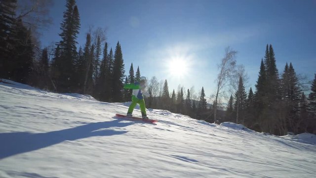 Sportsman in bright green clothes is jumping on board upon a slope of the hill at sunny winter day