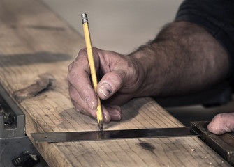 Closeup of carpenter's rough hands using a pencil and old square to mark a line on a wood board to cut using a chop saw