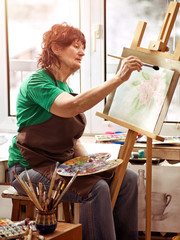 Obraz premium Artist painting easel in studio. Authentic senior woman has small home own business paints with brush in morning spring sunlight on window work place background.