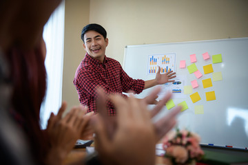 Handsome young man standing near whiteboard and pointing on the chart presentation perfect planning business with people group clapping.