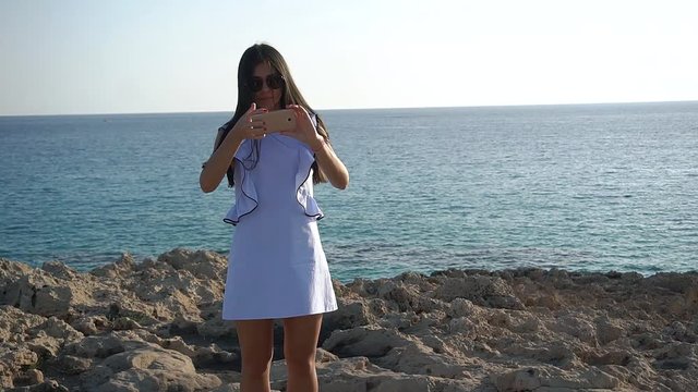 Young brunette woman in little blue dress taking a selfie at the background of rocks and sea.