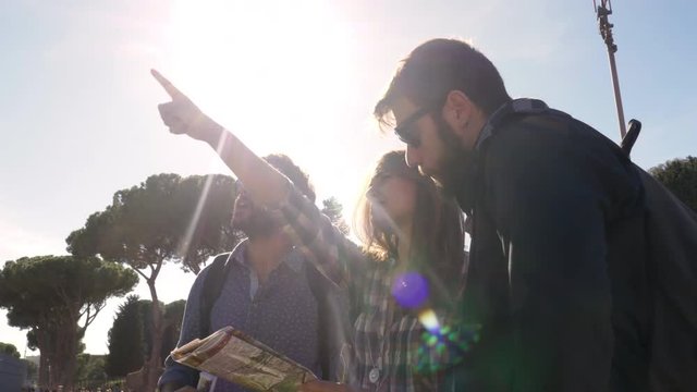 Three young friends tourists standing in front of colosseum in rome reading map guide for directions pointing with backpacks sunglasses happy beautiful girl long hair steadycam camera spinning around