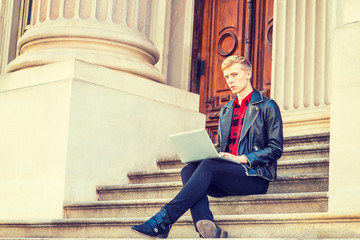 Young blonde American Man studying, working in New York, wearing black leather jacket, pants, leather shoes, sitting on stairs in office building on campus, reading, working on laptop computer..