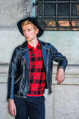 Young Man Casual Spring/Autumn Fashion in New York, wearing black leather jacket, patterned red undershirt, black hat, standing by wall on street, looking away. .