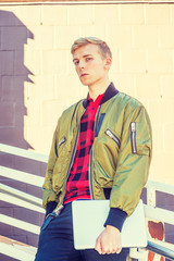 Young blonde American college student wearing green jacket unzipped, red patterned shirt, holding laptop computer, standing against railing by wall on campus in New York, relaxing, thinking..