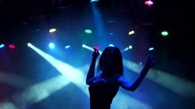 Silhouette of a slender dancing girl in a nightclub. Sexy girl dances in the dark with the light of multi-colored searchlights, slow movement.