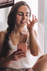 beautiful young woman sitting on the windowsill listen to music hold a smartphone