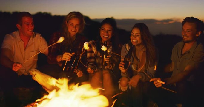 Group of friends relaxing around campfire roasting marshmallows, sunset bonfire lifestyle