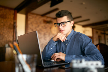 Thoughtful young caucasian businessman in glasses working on laptop computer. Pensive attractive hipster freelancer thinks over the solution of the problem with computer in cafe. Business concept.