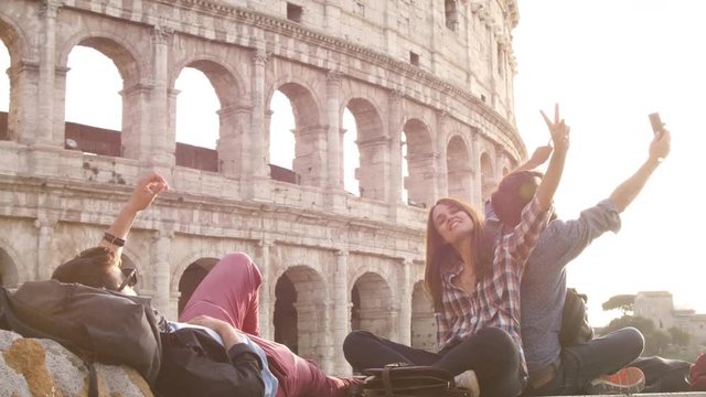 Three young friends tourists sitting in front of colosseum in rome at sunset taking selfies with smartphone with backpacks sunglasses happy beautiful girl long hair slow motion