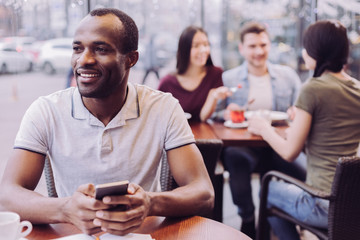 Funny answer. Energetic hopeful pensive man holding phone while staring aside and posing at cafe