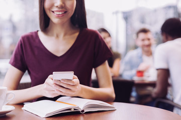 Technological advantage. Close up of tender young female hands using phone while woman smiling and notebook lying on the table